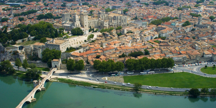 Vue aérienne du centre historique d'Avignon, avec sa majestueuse cathédrale, son pont en pierre enjambant le fleuve et ses bâtiments aux toits en terre cuite, serrés les uns contre les autres et entourés d'une végétation luxuriante.