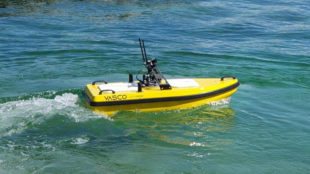 Le Bathydrone VASCO, un véhicule de surface sans pilote compact et jaune équipé d'antennes, glisse sur des eaux bleues-vertes claires, laissant de douces ondulations dans son sillage.