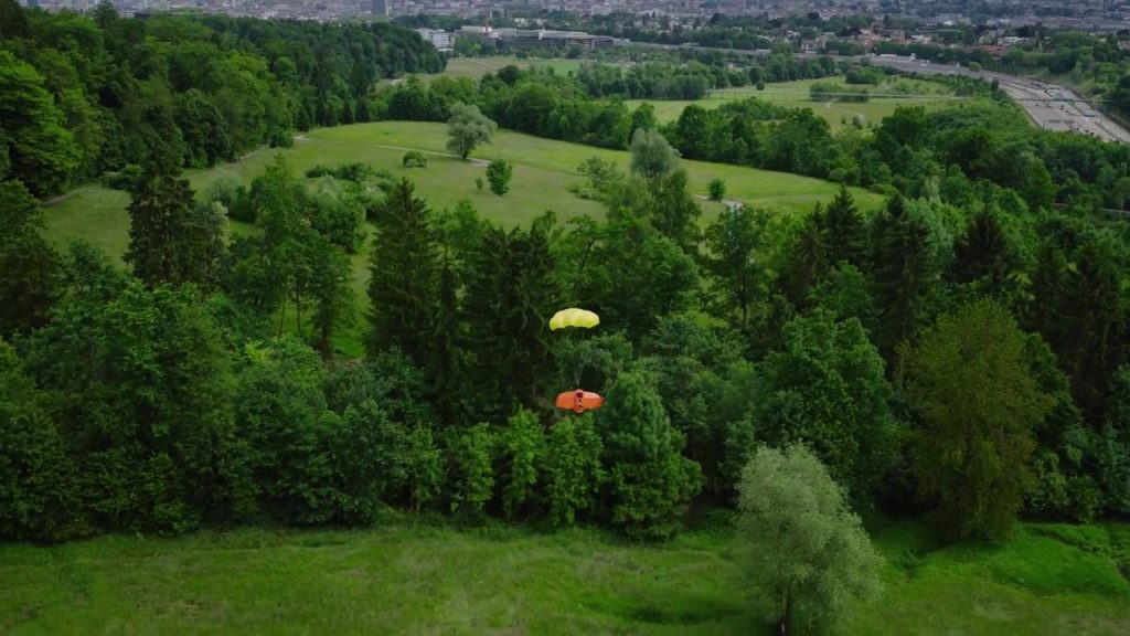 Une personne munie d'un parachute jaune descend au-dessus d'un paysage verdoyant tandis que le drone Aile Volante WingtraRAY scrute la scène, avec une ville lointaine visible en arrière-plan.