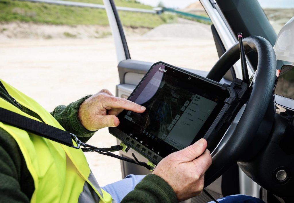 Un ouvrier vêtu d'un gilet de sécurité jaune utilise une tablette tactile montée sur le volant d'un véhicule, affichant un plan de chantier intégré à Aile Volante WingtraRAY. La porte ouverte du véhicule montre un chantier en cours à l'extérieur.
