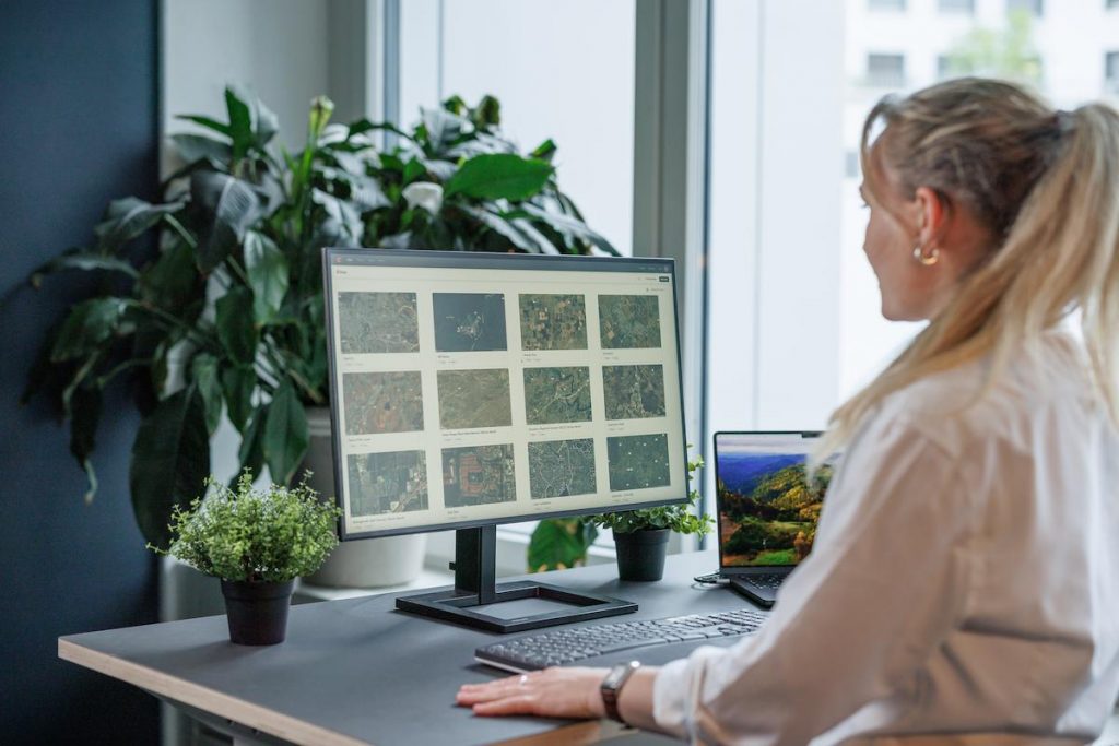 Une femme est assise à un bureau et regarde les images aériennes capturées par le drone Aile Volante WingtraRAY sur un grand écran d'ordinateur. Son bureau est décoré de plantes en pot et la lumière naturelle pénètre par une fenêtre voisine.