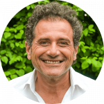 Un homme d'âge moyen aux cheveux noirs bouclés et au large sourire porte une chemise blanche et se tient en plein air devant un feuillage vert.