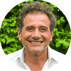 Un homme d'âge moyen aux cheveux noirs bouclés et au large sourire porte une chemise blanche et se tient en plein air devant un feuillage vert.