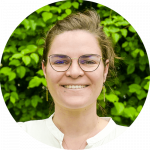 Une personne à la peau claire, portant des lunettes et une chemise blanche, sourit devant un fond de plantes à feuilles vertes.