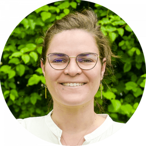 Une personne à la peau claire, portant des lunettes et une chemise blanche, sourit devant un fond de plantes à feuilles vertes.