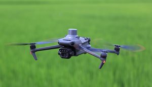 Le DJI Mavic 3 Multispectral, un drone gris équipé d'une caméra et de capteurs, plane au-dessus d'un champ vert flou, avec des hélices qui tournent et de petites lumières orange qui brillent sur ses bras.