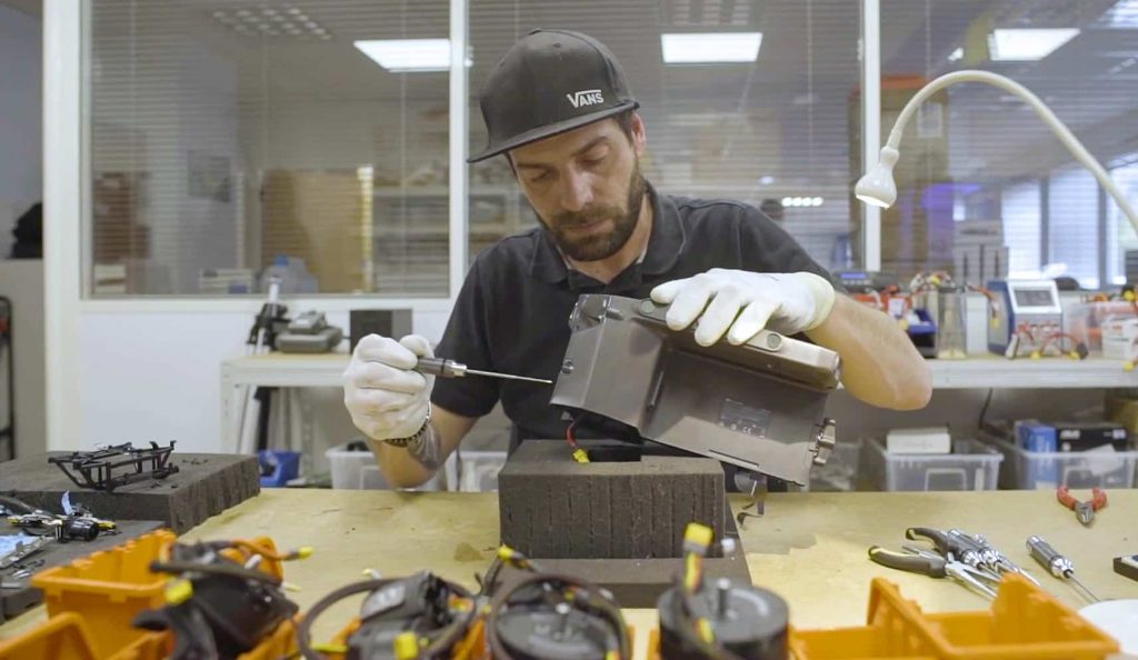 Un homme portant une casquette et des gants blancs utilise un tournevis pour assembler ou réparer un appareil électronique sur un établi, entouré d'outils et de composants, dans le cadre d'un atelier de réparation d'un centre actif.