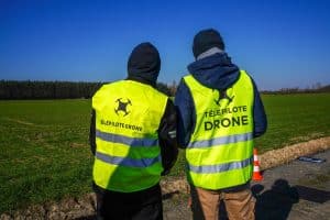 Deux personnes portant des gilets jaunes portant l'inscription « Telepilote Drone » se tiennent au bord d'une route, près d'un champ herbeux, avec en arrière-plan un ciel bleu éclatant et des arbres. Cette scène met en évidence la présence de télépilotes certifiés pilotant des drones dans un cadre naturel et à ciel ouvert.