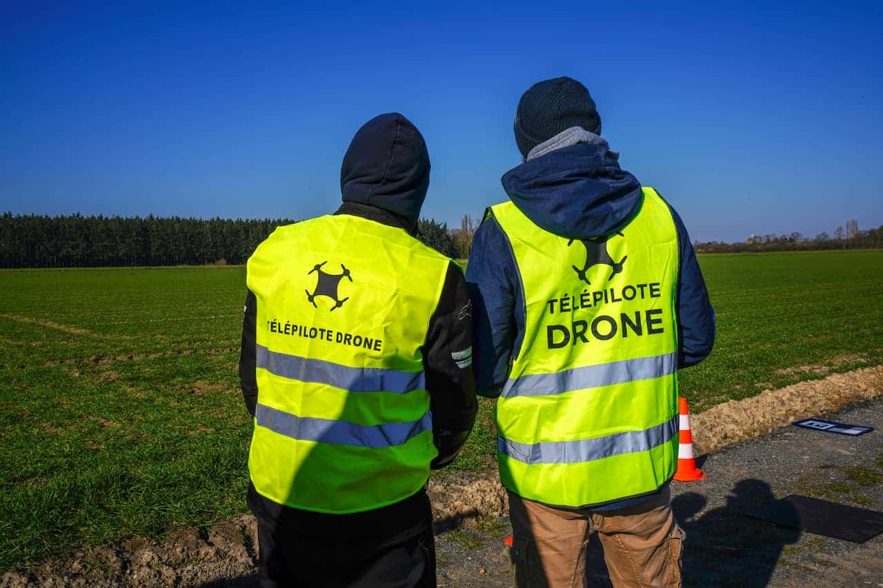 Deux personnes portant des gilets jaunes portant l'inscription « Telepilote Drone » se tiennent au bord d'une route, près d'un champ herbeux, avec en arrière-plan un ciel bleu éclatant et des arbres. Cette scène met en évidence la présence de télépilotes certifiés pilotant des drones dans un cadre naturel et à ciel ouvert.