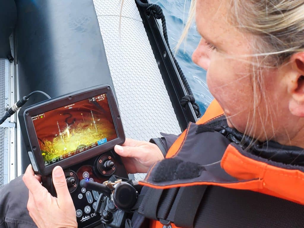 Une personne vêtue d'une veste orange est assise dans un bateau ; elle manipule un écran portable sur lequel s'affiche une scène sous-marine, probablement pour piloter un véhicule télécommandé ou un drone sous-marin.