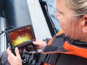 Une personne vêtue d'une veste orange est assise dans un bateau ; elle manipule un écran portable sur lequel s'affiche une scène sous-marine, probablement pour piloter un véhicule télécommandé ou un drone sous-marin.