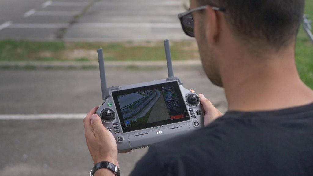 Une personne tenant et utilisant une télécommande de drone DJI avec un écran d'affichage montrant une vue aérienne en direct, debout en plein air près d'un parking vide.