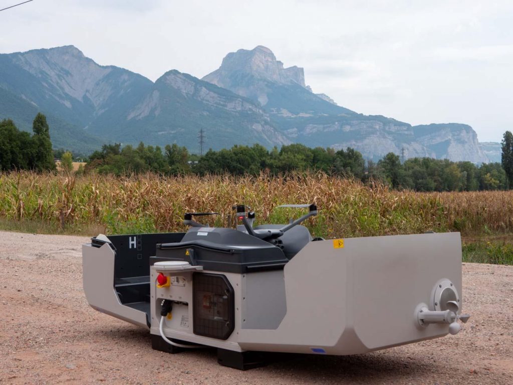 Une machine industrielle moderne et grise se trouve sur une route de gravier près d'un champ de maïs, avec des arbres verts et de hautes montagnes en arrière-plan sous un ciel nuageux.