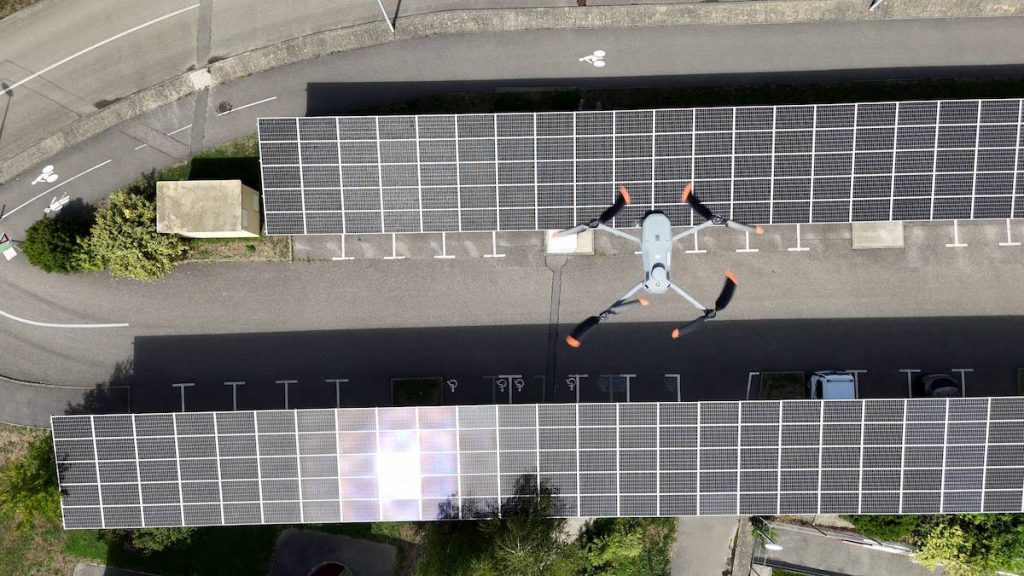 Un drone survole deux parkings couverts dont les toits sont équipés de panneaux solaires, vus d'une vue aérienne lors d'une journée ensoleillée.