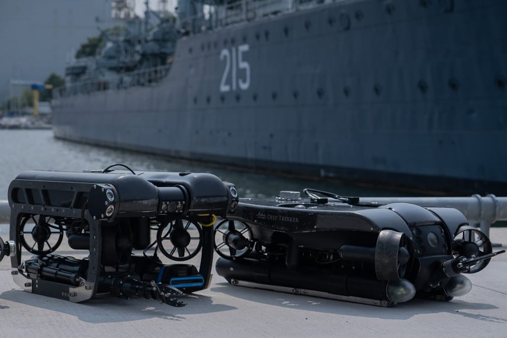 Deux drones sous-marins sont assis sur un quai devant un navire de guerre dont la coque porte le numéro 215. La scène semble se dérouler dans un port.