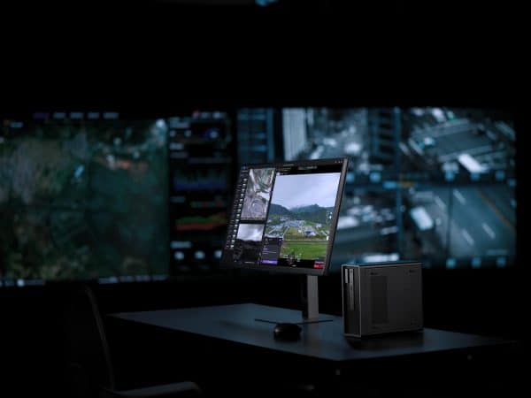 Dans une pièce sombre, un bureau est équipé d'un moniteur et d'un ordinateur de bureau fonctionnant avec DJI Flighthub 2 On-Premises, affichant des séquences de surveillance ; on peut voir en arrière-plan de grands écrans flous avec des flux de sécurité.