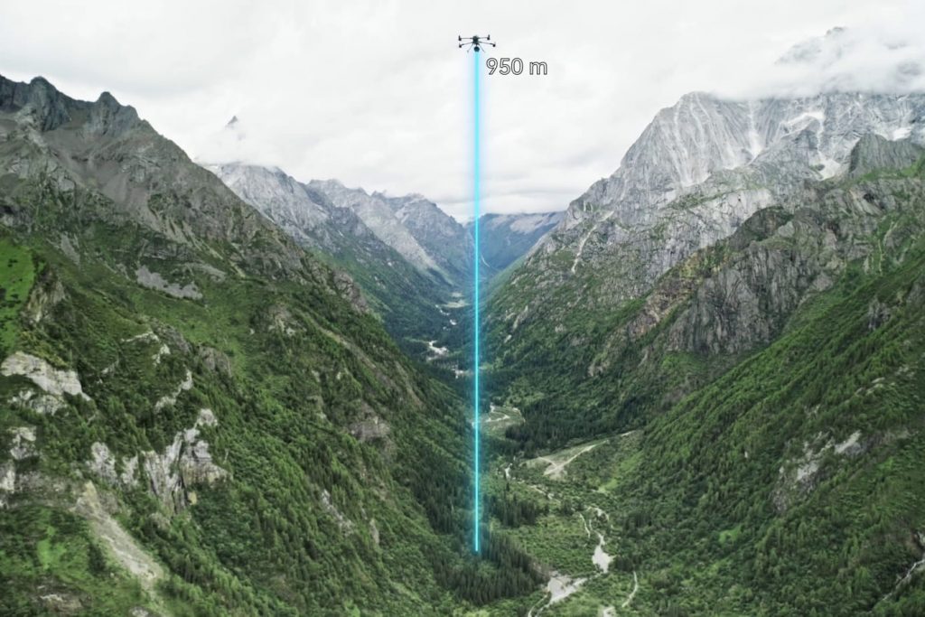 Un drone DJI L3 LiDAR plane à 950 mètres au-dessus d'une vallée montagneuse verdoyante, émettant un faisceau bleu vertical alors que des sommets escarpés et couverts de nuages s'élèvent à l'arrière-plan.