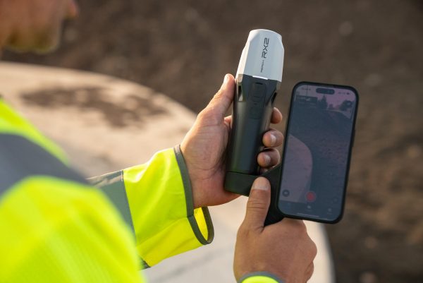 Une personne portant une veste de haute visibilité tient un appareil portable Emlid Reach RX2 dans une main et un smartphone avec une application de caméra ouverte dans l'autre, à l'extérieur par une journée ensoleillée.