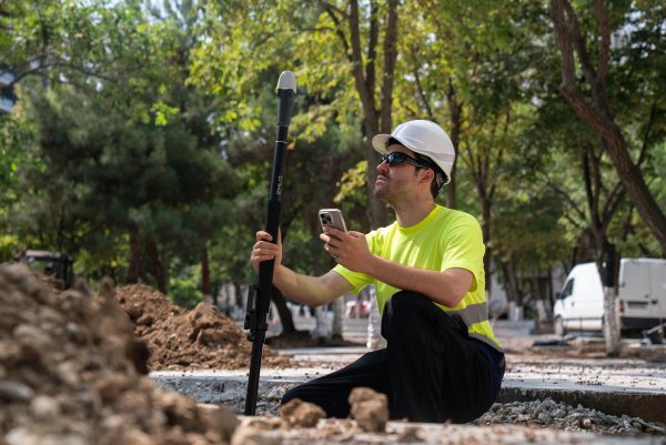 Un ouvrier du bâtiment portant un casque blanc et une chemise jaune est agenouillé à l'extérieur, tenant une canne d'arpentage Emlid Reach RX2 et un smartphone, entouré de terre et d'arbres par une journée ensoleillée.