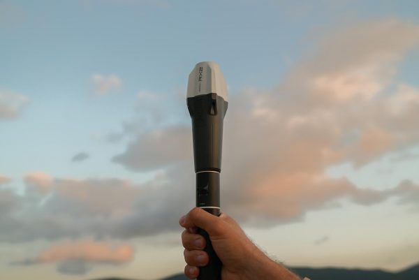 Une main tient l'Emlid Reach RX2 dans un ciel de coucher de soleil avec de doux nuages, avec des collines lointaines visibles au bas de l'image.