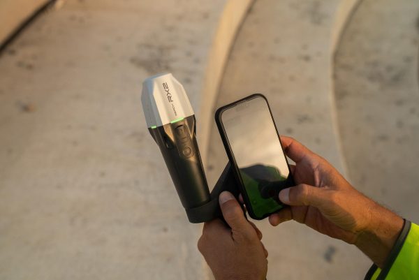 Une personne tient un appareil Emlid Reach RX2 attaché à un smartphone pendant qu'elle l'utilise à l'extérieur, avec des marches en béton incurvées visibles à l'arrière-plan.