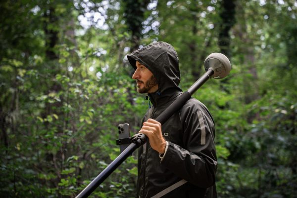 Une personne vêtue d'une veste de pluie sombre et d'une capuche se tient dans une forêt verdoyante, tenant une grande perche surmontée de l'Emlid Reach RS4, un appareil sphérique probablement utilisé pour l'arpentage ou la cartographie.