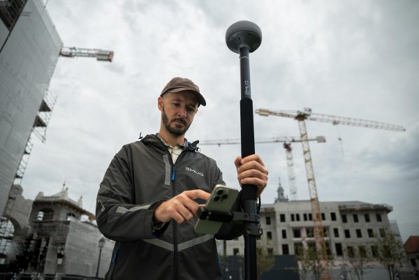 Un homme vêtu d'une veste et d'une casquette utilise l'appareil de topographie GPS Reach RS4 d'Emlid avec son smartphone sur un chantier de construction, où des grues et une structure partiellement construite se dressent sous un ciel nuageux.