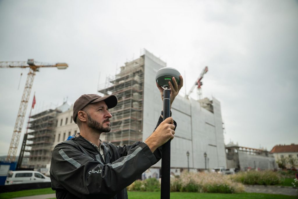 Un homme utilise l'équipement topographique Emlid Reach RS4 à l'extérieur, près d'un chantier de construction, avec des grues et des échafaudages visibles sur plusieurs grands bâtiments à l'arrière-plan.