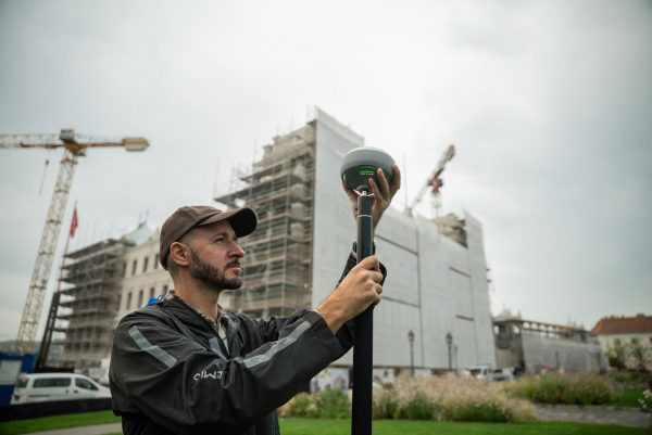 Un homme utilise l'équipement topographique Emlid Reach RS4 à l'extérieur, près d'un chantier de construction, avec des grues et des échafaudages visibles sur plusieurs grands bâtiments à l'arrière-plan.