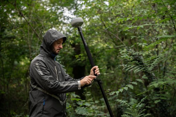 Une personne vêtue d'une veste de pluie à capuche ajuste un appareil GPS Reach RS4 d'Emlid monté sur un poteau dans une forêt dense et verdoyante.