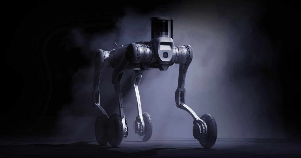 Un robot Unitree B2-W à quatre pattes avec des roues sur chaque jambe se tient dans un environnement dramatique et enfumé, éclairé par un projecteur sur un fond sombre.