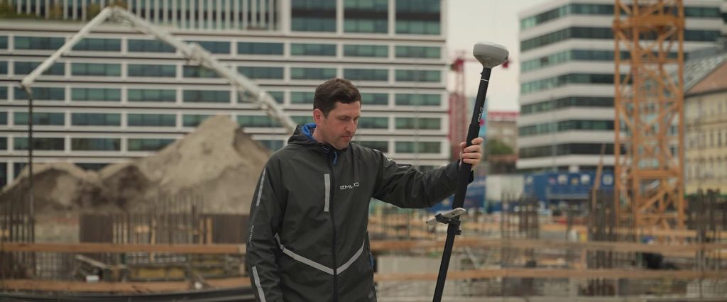 Un homme vêtu d'une veste de pluie tient une canne GPS Emlid Reach RS4 Pro sur un chantier de construction, avec en arrière-plan des immeubles de bureaux modernes, des grues et des tas de terre.