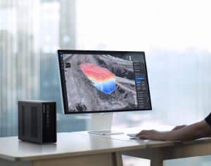 Une personne utilise DJI Flighthub 2 On-Premises sur un ordinateur de bureau pour visualiser une carte en 3D avec des dégradés de couleurs et des superpositions de données dans un bureau avec de grandes baies vitrées. Une tour d'ordinateur noire se trouve à côté de l'écran.
