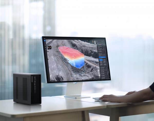 Une personne utilise DJI Flighthub 2 On-Premises sur un ordinateur de bureau pour visualiser une carte en 3D avec des dégradés de couleurs et des superpositions de données dans un bureau avec de grandes baies vitrées. Une tour d'ordinateur noire se trouve à côté de l'écran.