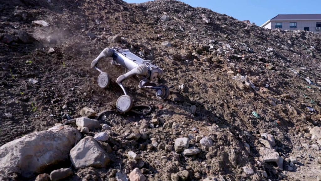 Un robot Unitree B2-W à quatre pattes grimpe une colline rocailleuse et couverte de terre sous un ciel bleu clair, avec un bâtiment en arrière-plan.