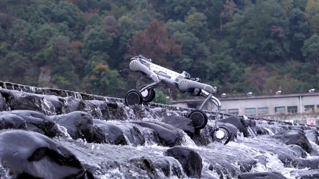 Le rover robotique Unitree B2-W à quatre roues escalade des rochers humides et irréguliers près d'un ruisseau, avec une colline boisée et un bâtiment en arrière-plan.