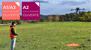 Une personne portant un gilet de sécurité orange pilote un drone dans un champ herbeux. Deux étiquettes colorées en français indiquent les sous-catégories de drones A1/A3 et A2. Un tapis d'héliport orange est au sol. Des arbres et des collines se trouvent à l'arrière-plan.