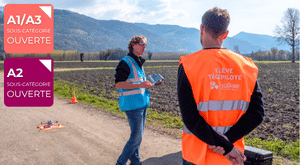 Deux personnes se tiennent sur une route de campagne ; l'une d'elles porte un gilet portant l'inscription « ÉLÈVE TÉLÉPILOTE ». Du matériel de pilotage de drone est posé au sol. Des étiquettes indiquant les catégories de drones A1/A3 et A2 apparaissent à gauche. Des champs et des montagnes sont visibles en arrière-plan.