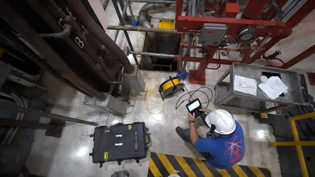 Un travailleur portant un casque blanc et une chemise bleue utilise un appareil portatif à proximité d'équipements industriels, de câbles et d'équipements de sécurité dans une installation. Des bandes de danger noires et jaunes marquent le sol près d'une fosse ouverte.