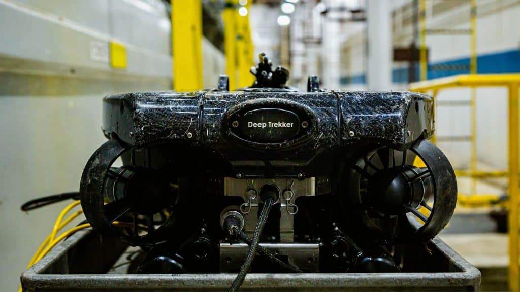 Vue rapprochée d'un véhicule sous-marin télécommandé (ROV) de couleur noire, appelé Deep Trekker, placé sur un chariot métallique dans un environnement industriel, avec des garde-corps et des équipements jaunes visibles en arrière-plan.