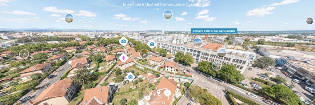 Vue aérienne d'une ville avec des maisons résidentielles, des jardins verdoyants et de grands bâtiments industriels ; divers points de repère sont indiqués par des marqueurs bleus et des bulles de texte, sous un ciel dégagé.