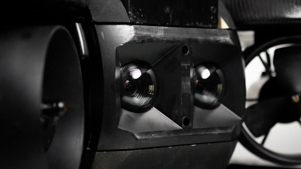 Gros plan de deux objectifs de caméra grand angle montés côte à côte sur le Deep Trekker Spectra, un dispositif noir à pivot angulaire, peut-être destiné à une utilisation robotique ou à un drone, avec des composants mécaniques flous visibles à l'arrière-plan.