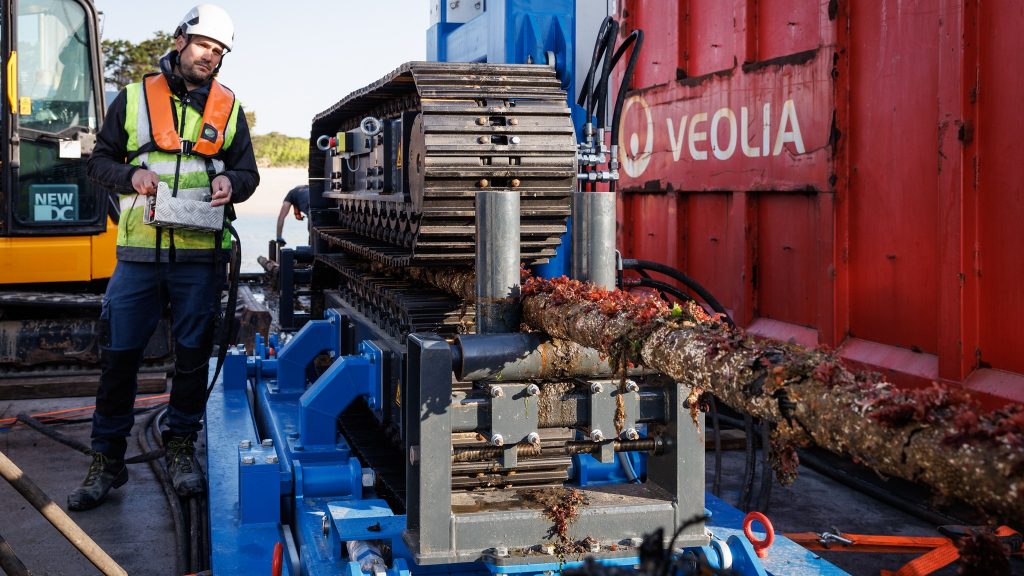 Un ouvrier en tenue de sécurité utilise une machine pour enlever les algues d'un câble épais et couvert de balanes. La scène se déroule en plein air, près d'un conteneur rouge portant l'inscription Veolia et d'un engin de chantier.