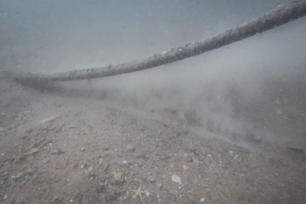 Un long tuyau sombre gît sous l'eau sur le lit d'une rivière trouble et rocailleuse. Des sédiments tourbillonnent visiblement autour de lui, créant un effet de trouble dans l'eau.
