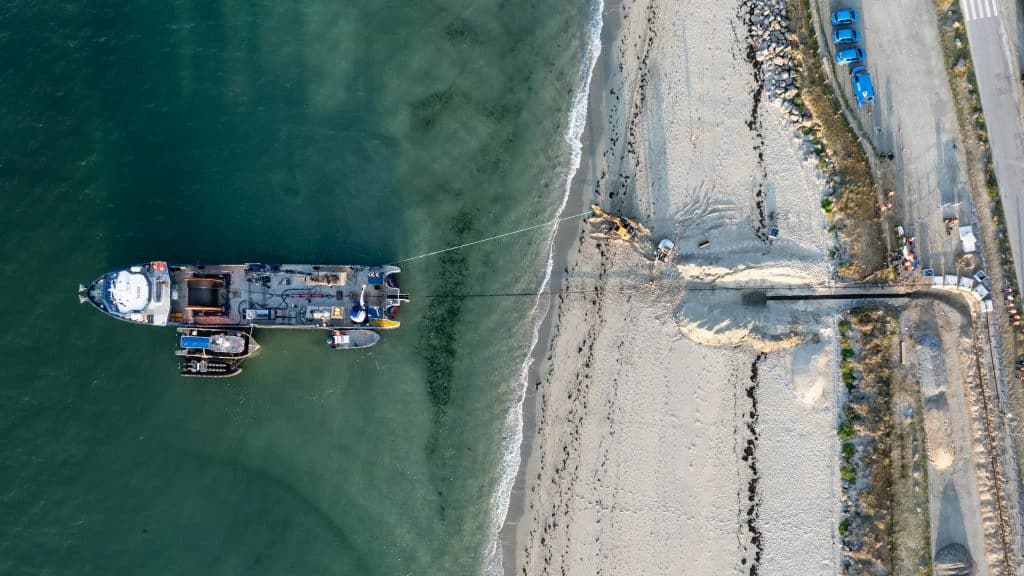 Vue aérienne d'un navire ancré près du rivage avec plusieurs petits bateaux à proximité. Un grand tuyau s'étend du navire jusqu'à une plage de sable, où des engins de chantier travaillent près de voitures garées et d'une zone rocheuse.