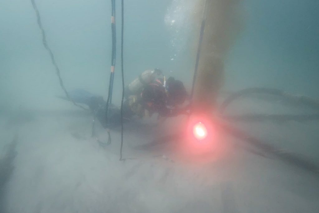 Un plongeur sous l'eau actionne une machine, remuant du sable et des sédiments, avec une lumière rouge vive éclairant la scène et plusieurs câbles ou tuyaux visibles autour de lui.