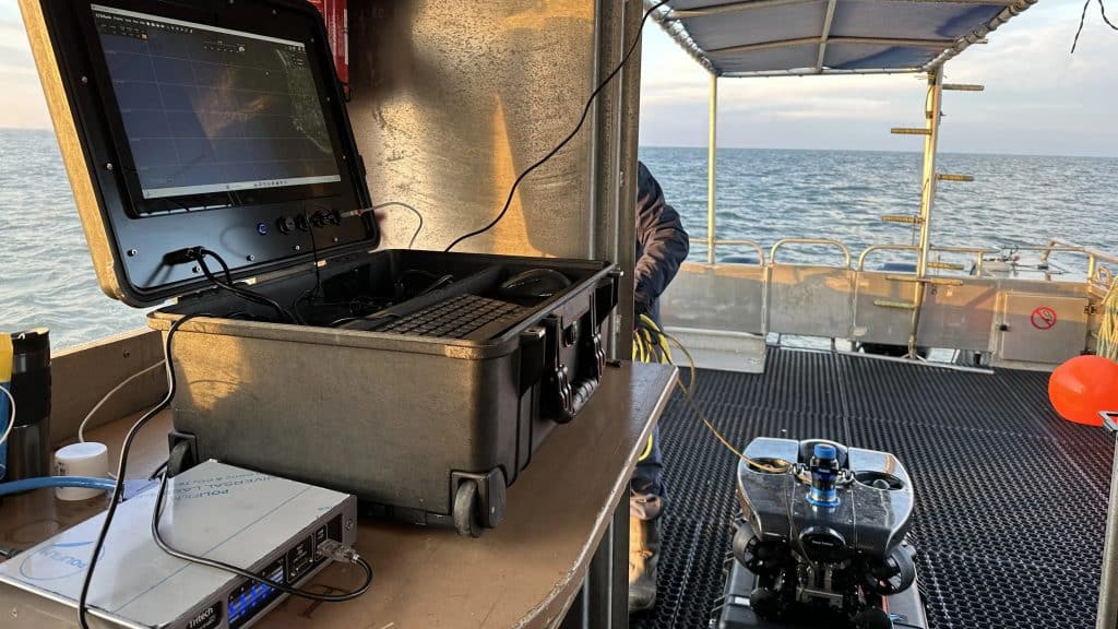 Un ordinateur portable robuste affichant des données sonar est posé sur une table métallique à bord d'un navire de recherche, avec des câbles et du matériel à proximité. Le bateau est en pleine mer, sous un ciel partiellement nuageux.
