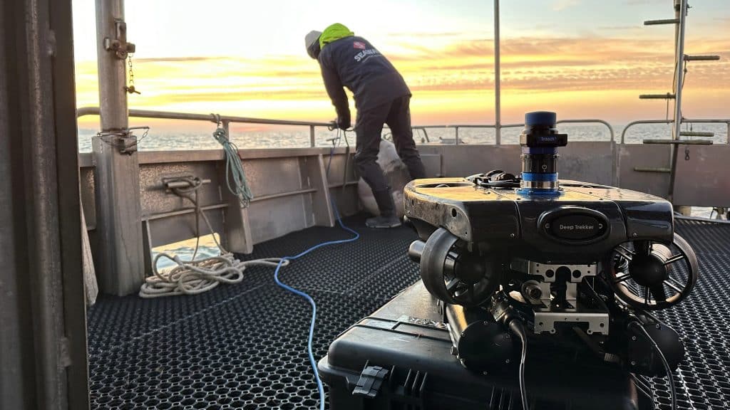 Une personne en tenue de plein air se tient sur un bateau au coucher du soleil, manipulant une corde. Au premier plan, un véhicule sous-marin télécommandé (ROV) appelé Deep Trekker repose sur une caisse.