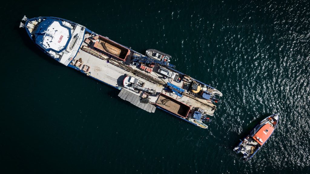 Vue aérienne d'un grand cargo chargé de conteneurs et de véhicules, flottant sur des eaux sombres, avec un plus petit bateau orange et blanc positionné à proximité sur la droite.