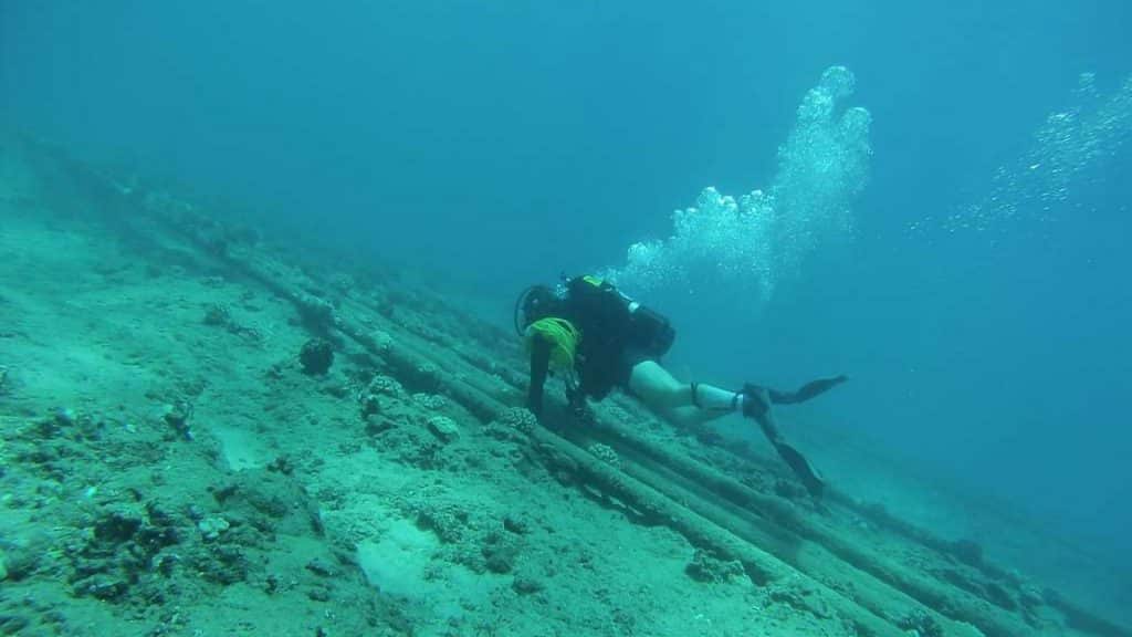 Un plongeur sous-marin en tenue complète nage près de tuyaux ou de câbles sous-marins parallèles au fond de l'océan, entouré de rochers épars et d'animaux marins, avec des bulles remontant à la surface.